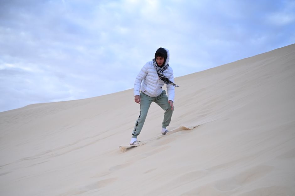 A man in a white jacket sandboarding on the vast desert sand dunes under a cloudy sky.